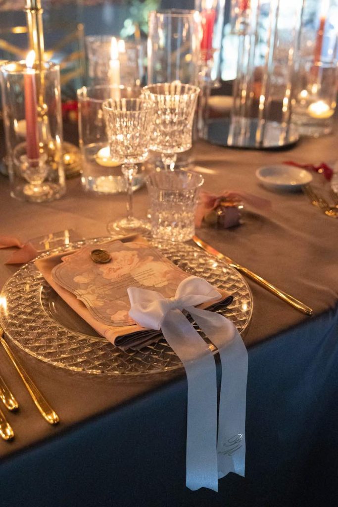 In questa foto angolare, un dettaglio della mise en place del matrimonio di Maria Pia e PierPaolo con un sottopiatto trasparente, posate d'argento, bicchieri in vetro trasparente, tovagliolo rosa, menù con stampa floreale e un nastro di colore bianco