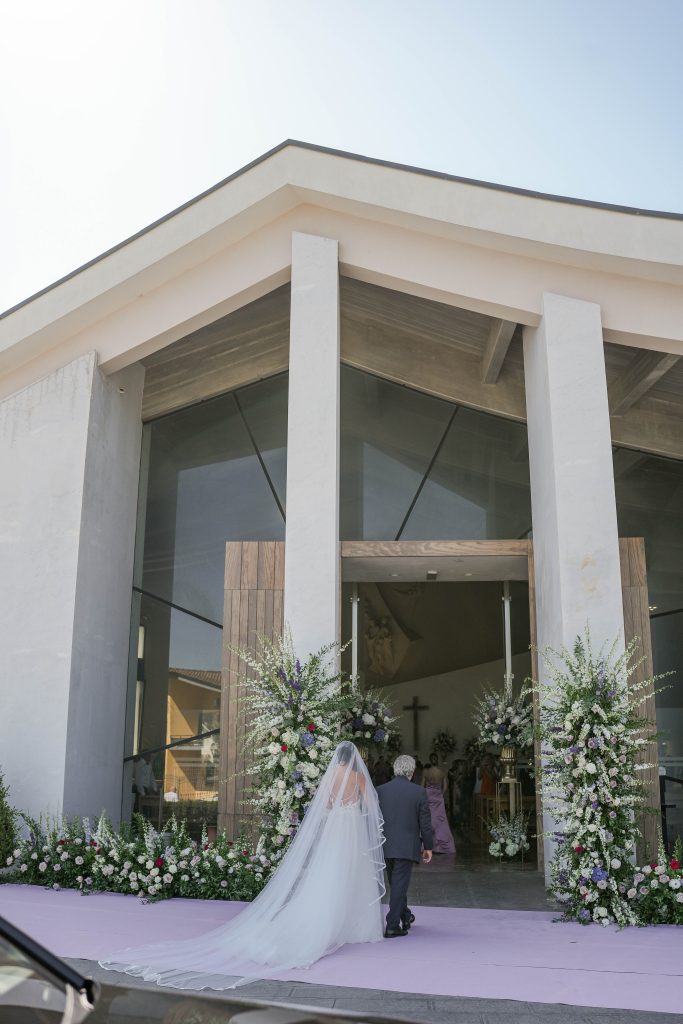 In questa foto, una sposa di spalle, con un lungo velo bianco, mentre entra in chiesa accompagnata da un uomo anziano. La chiesa ha un’architettura moderna, con facciata chiara e linee geometriche pulite. L’ingresso è incorniciato da grandi composizioni floreali di colore bianco, viola e rosa, disposte su entrambi i lati della porta e lungo il percorso d’ingresso.