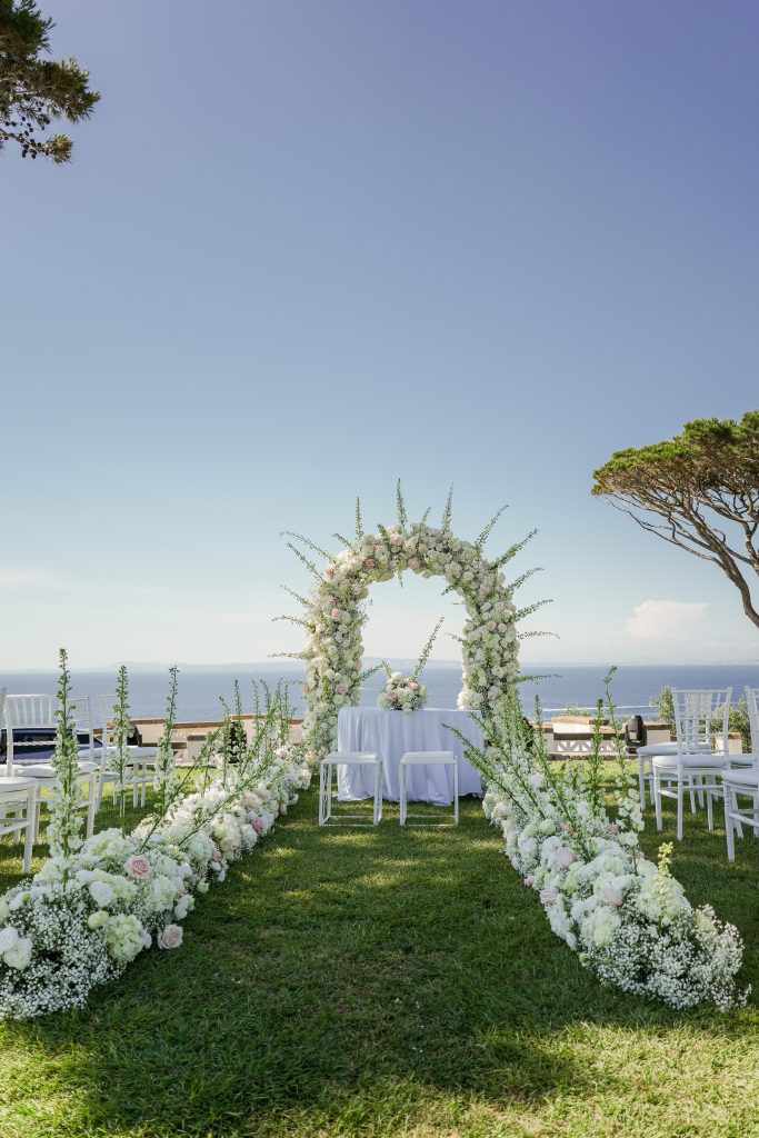 In questa foto si vede una scena di uno dei matrimoni di Stefano Miranda un arco floreale di colore bianco con sfumature di rosa, collocato al centro di uno spazio erboso con vista mare. Davanti all’arco si trova un tavolo con tovaglia bianca e due sgabelli, mentre tutto intorno ci sono sedie bianche disposte ordinatamente per la cerimonia. Il cielo è limpido e azzurro, e si intravedono pini marittimi sul lato.