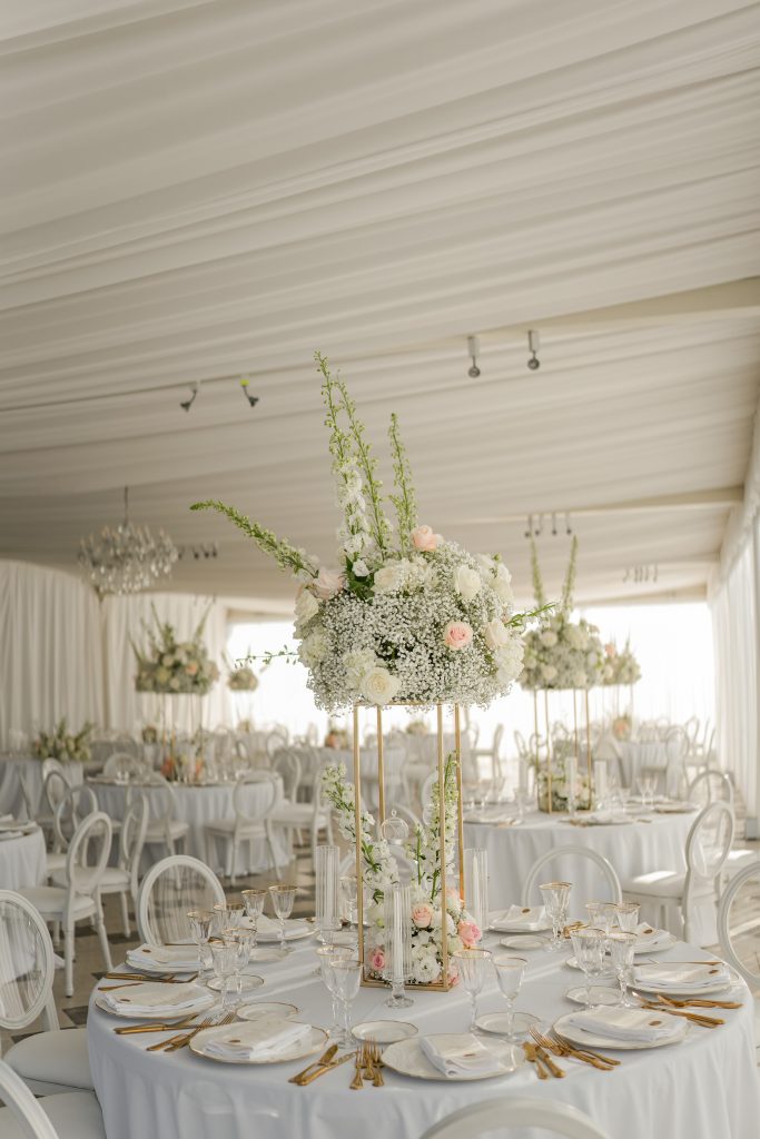 In questa foto si vede una scena di uno dei matrimoni di Stefano Miranda un tavolo apparecchiato con bicchieri e piatti in cristallo, circondato da sedie di colore bianco. Il centrotavola è molto scenografico, composto da un grande bouquet di fiori di colore fucsia, viola, rosso e oro, con orchidee che scendono a cascata. L’ambiente è illuminato da candele accese in cilindri di vetro trasparente, creando un’atmosfera romantica e intensa.