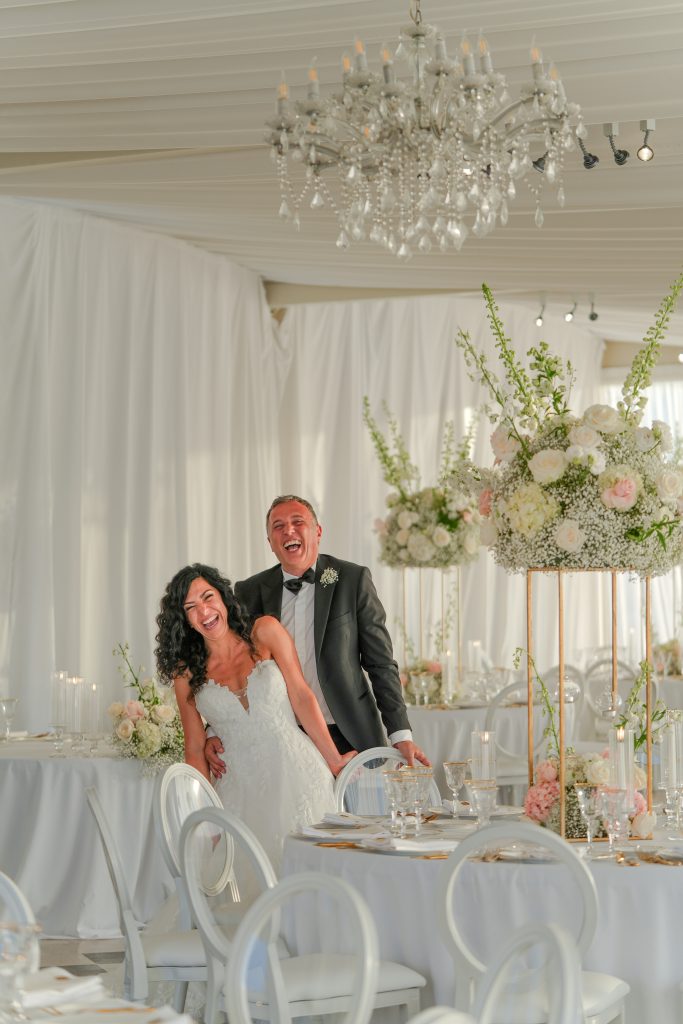 In questa foto si vede una scena di uno dei matrimoni di Stefano Miranda in cui una coppia di sposi sorride in camera in una elegante sala ricevimenti allestita con fiori colorati