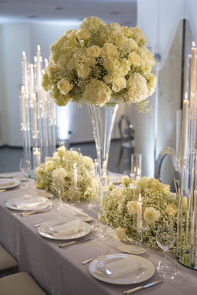 In questa foto un tavolo per matrimonio allestito da  Stefano Miranda con un centrotavola è composto da una grande composizione floreale di colore bianco e crema, con rose e ortensie, posta su un alto vaso trasparente. Attorno, candele alte in cilindri di vetro creano un’atmosfera raffinata. Il tovagliato è di colore chiaro e ogni posto è apparecchiato con piatti bianchi, posate in acciaio e calici trasparenti.