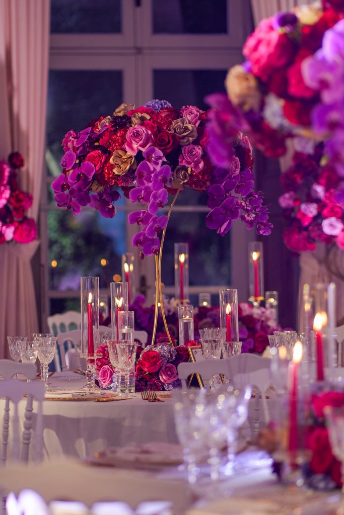 In questa foto la sala di un ricevimento di nozze allestita dal Wedding Planner Stefano Miranda con tavoli rotondi rivestiti da tovaglie di colore bianco. Al centro di ciascun tavolo, un alto centrotavola floreale con rose di colore bianco e rosa pallido, ortensie e gypsophila, sorretto da una struttura dorata. Le sedie sono di colore bianco, abbinate alla palette chiara dell’ambiente. L'atmosfera è luminosa e sofisticata, con drappeggi bianchi che ricoprono pareti e soffitto.