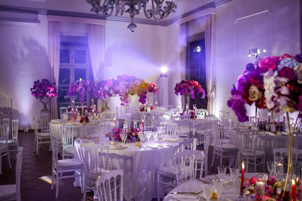 In questa foto si vede una scena di uno dei matrimoni di Stefano Miranda una sala elegante con numerosi tavoli rotondi decorati con fiori di colore fucsia, viola e rosso. Ogni tavolo è arricchito da un centrotavola alto e floreale e da candele sottili e luminose. Le pareti sono bianche, le tende rosa chiaro, e un gioco di luci colorate, tra cui il viola, dona un tocco moderno e scenografico all’ambiente.
