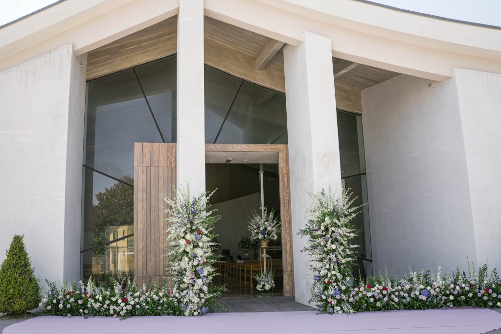 In questa foto la facciata esterna di una chiesa moderna, con una grande porta centrale in legno chiaro e due colonne bianche ai lati. L’ingresso è decorato con composizioni floreali di colore bianco, lilla, blu e rosso poste alla base e ai lati della porta, creando una scenografia elegante e romantica per l’arrivo degli sposi.