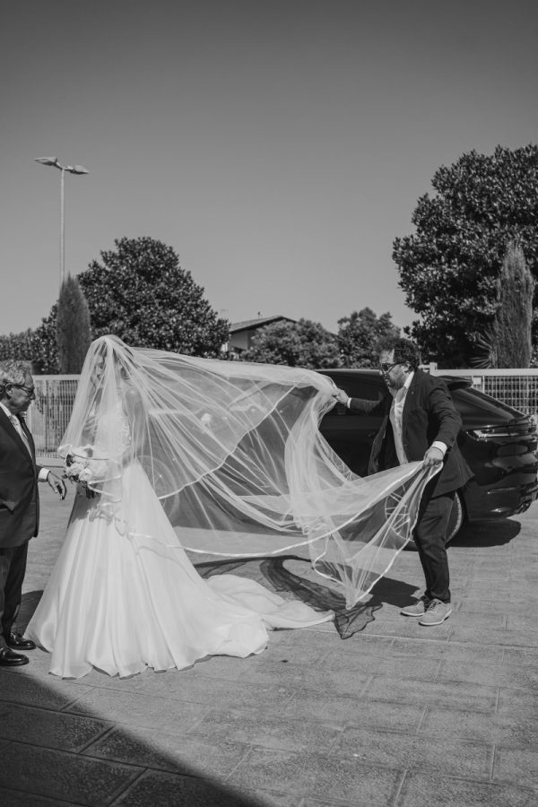 In questa foto in bianco e nero si vede una scena di uno dei matrimoni di Stefano Miranda  una sposa con un lungo velo di colore bianco che viene sistemato da un uomo mentre scende da un'auto scura. Indossa un abito nuziale ampio, anch’esso di colore bianco, e tiene in mano un piccolo bouquet. Accanto a lei, un altro uomo elegante l’attende. La scena è all’aperto, su una pavimentazione in pietra, con alberi e un cielo sereno sullo sfondo.