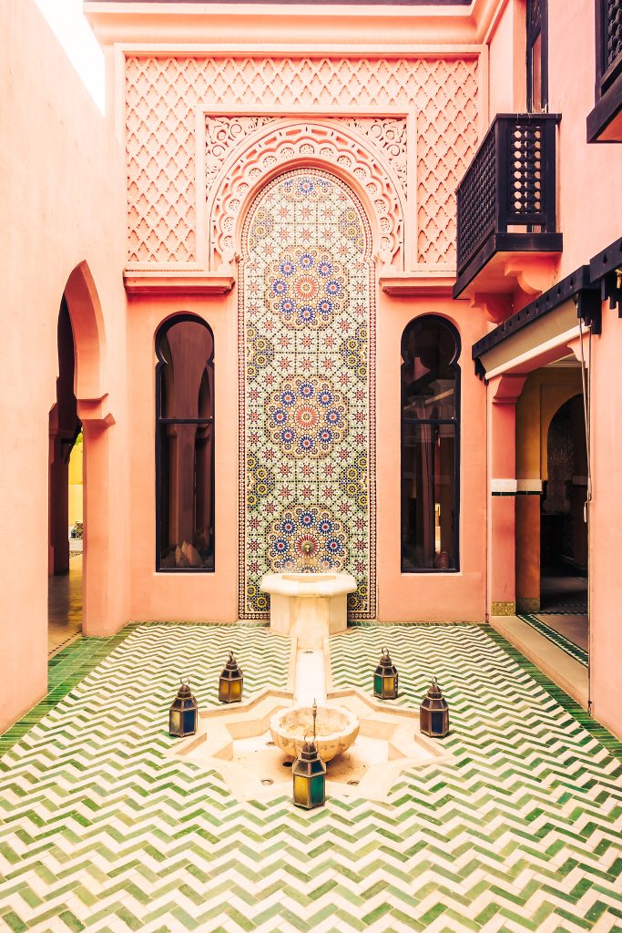 Cortile interno decorato con piastrelle geometriche verde-bianco, una fontana centrale circondata da lanterne, pareti rosa con archi e un grande mosaico ornamentale.