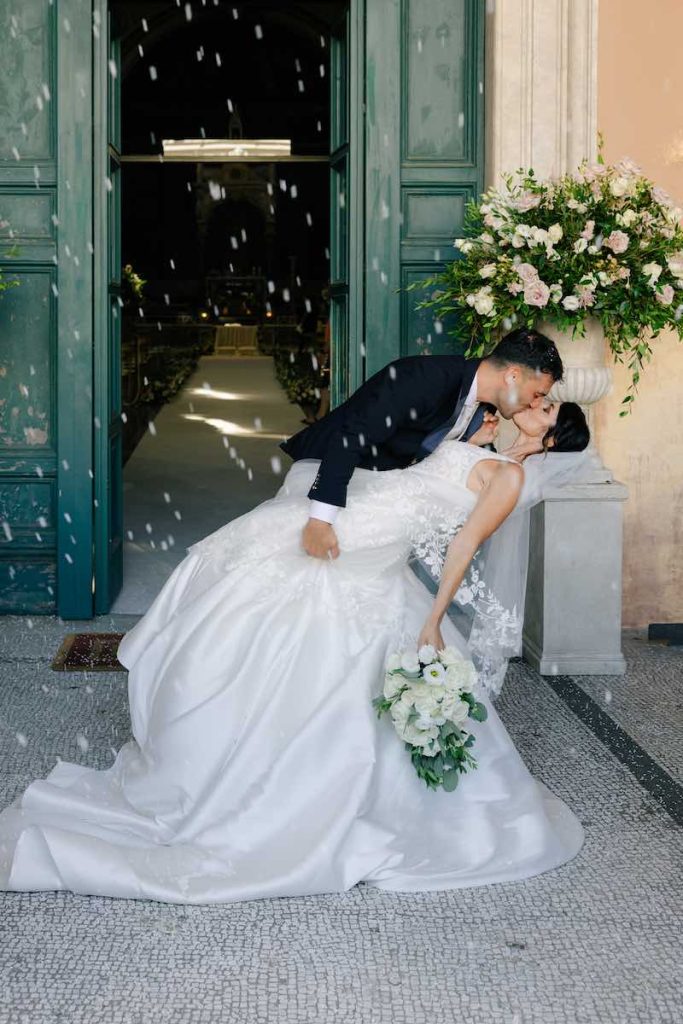In questa foto, due sposi che si baciano dopo la cerimonia di nozze mimando un casqué davanti all'ingresso di una chiesa. Lei tiene in mano un bouquet di fiori di colore bianco