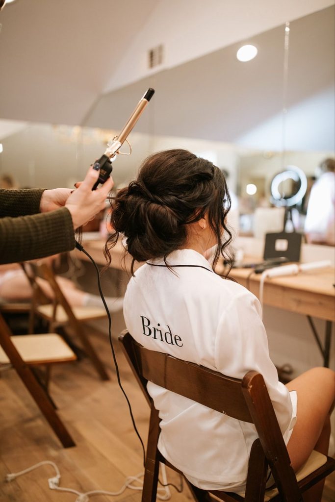 in questa foto si vede un'acconciatura sposa capelli lunghi raccolti di colore castano scuro. la sposa è seduta mentre un parrucchiere sposa ultima l'acconciatura