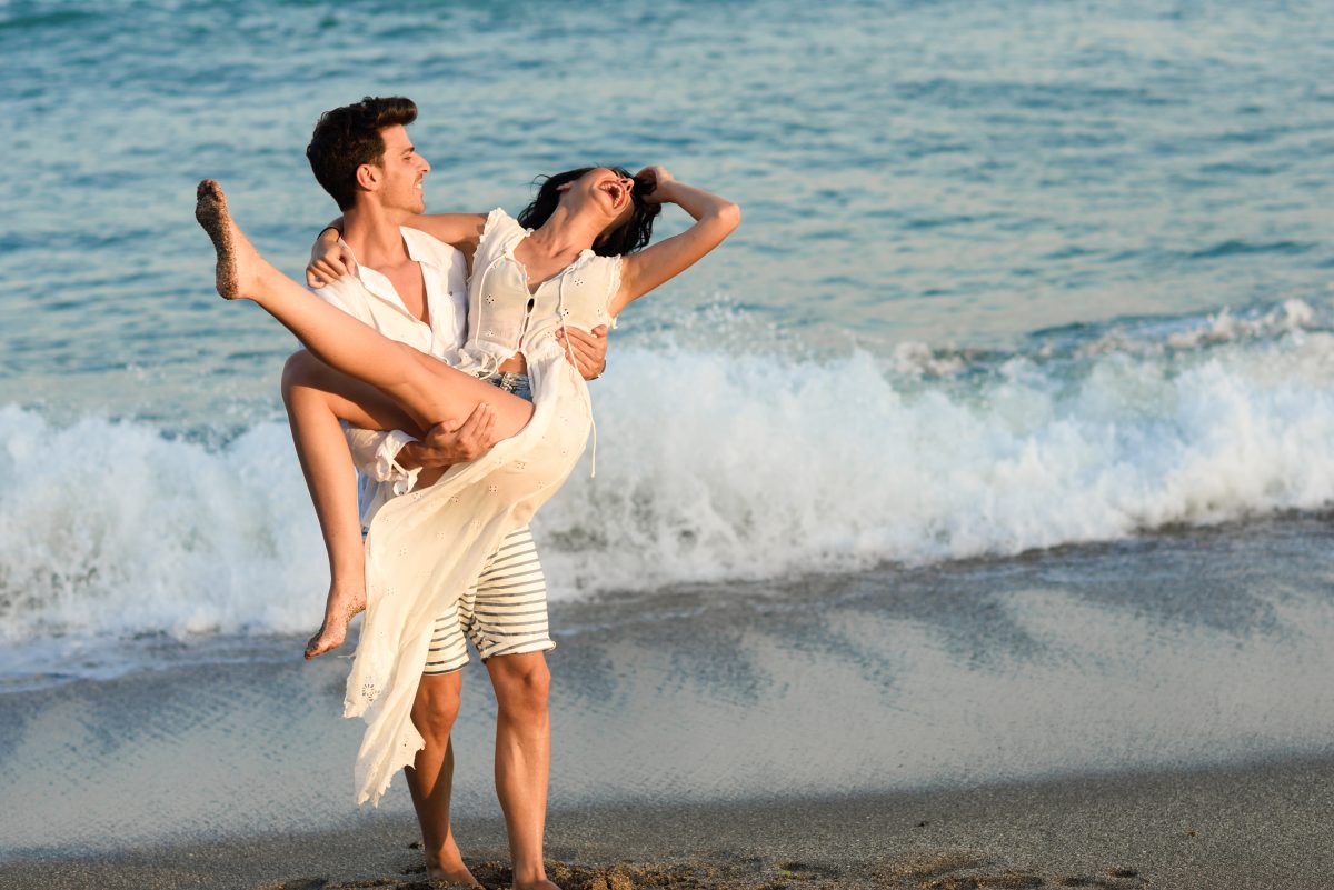 Giovane coppia felice che passeggia su una splendida spiaggia. Un uomo buffo porta una donna in braccio. Persone in abiti casual.
