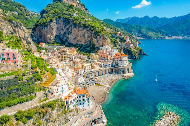 Veduta panoramica di una pittoresca cittadina italiana affacciata sul mare turchese, incastonata tra alte scogliere verdi. Le case colorate si arrampicano sulle colline, con una spiaggia sabbiosa e un porticciolo in basso