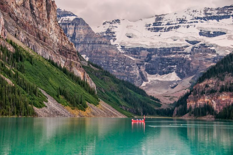 Lago glaciale dai toni verde smeraldo, incorniciato da alte montagne con vette innevate. Due canoe rosse solcano l’acqua tranquilla. Intorno solo foreste e natura selvaggia.