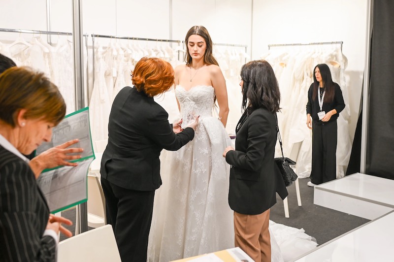 In questa foto, una modella indossa un abito da sposa mentre una stilista lo mostra ad una buyer