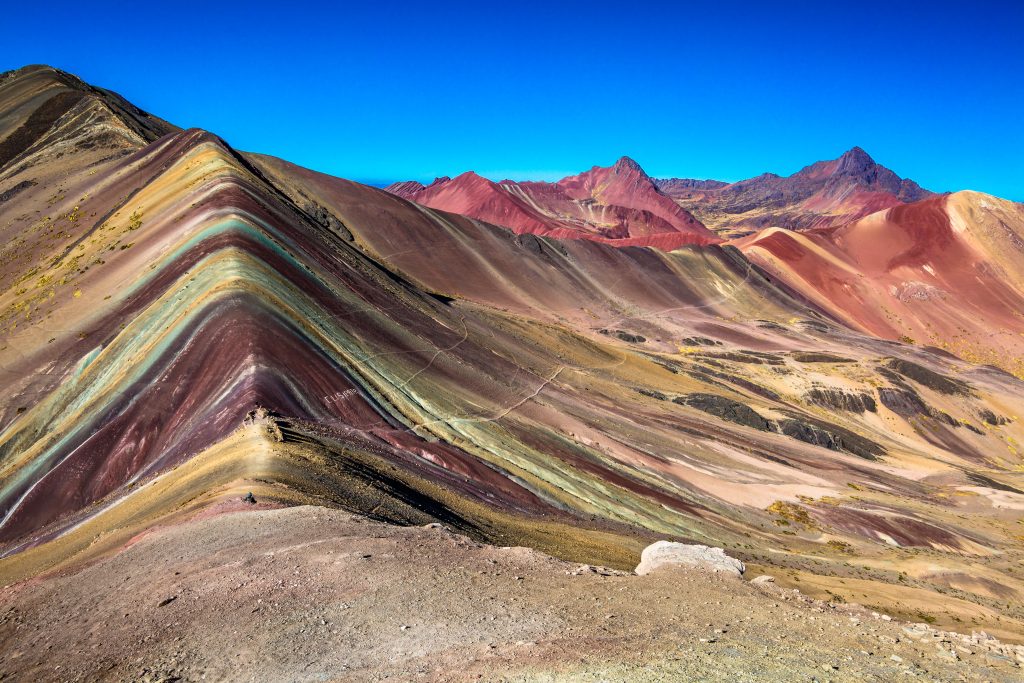 Vista panoramica di una montagna dalle striature naturali multicolore: tonalità di rosso, giallo, verde, azzurro e viola si susseguono lungo i fianchi della montagna, creando un effetto visivo sorprendente. Sullo sfondo, altre vette dai colori caldi si ergono sotto un cielo blu intenso e limpido.