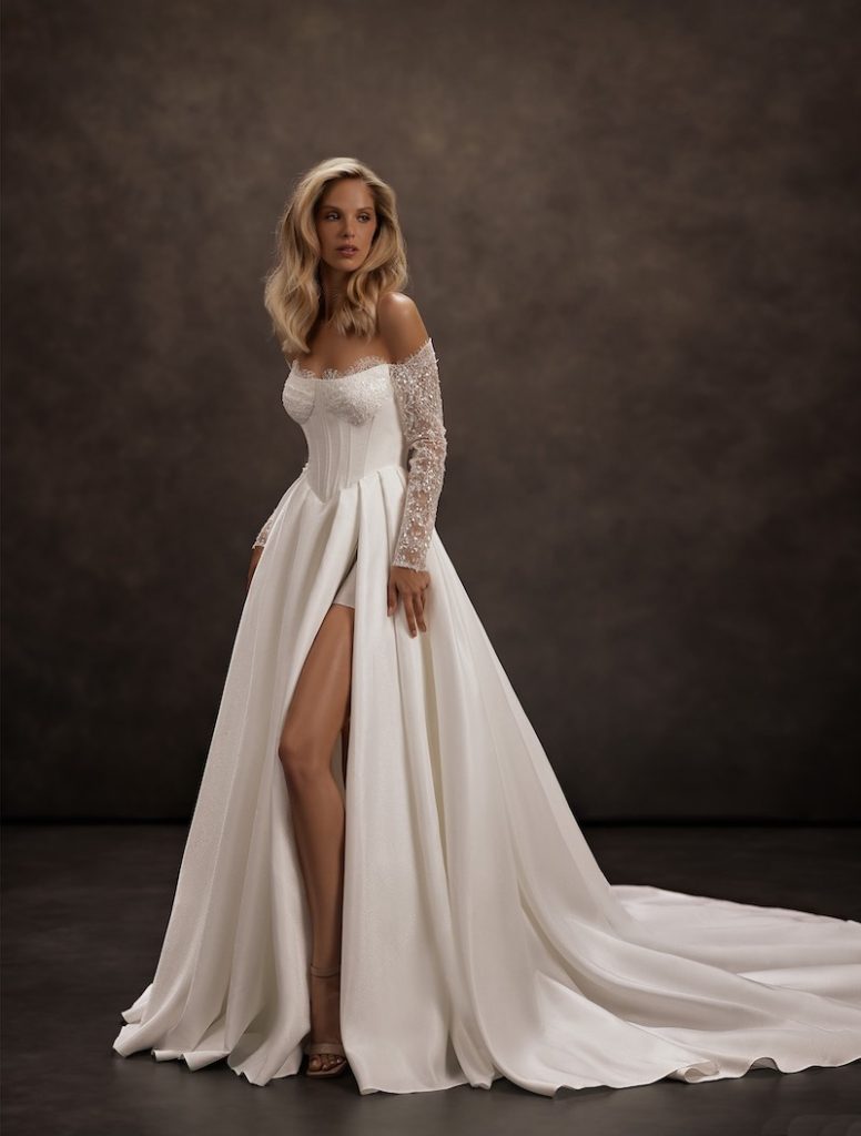 In questa foto, una modella posa  indossando un abito da sposa Jeorjett 2026 modello ad A in mikado di seta con spacco profondo laterale, bustier e maniche lunghe effetto illusion con ricami preziosi 