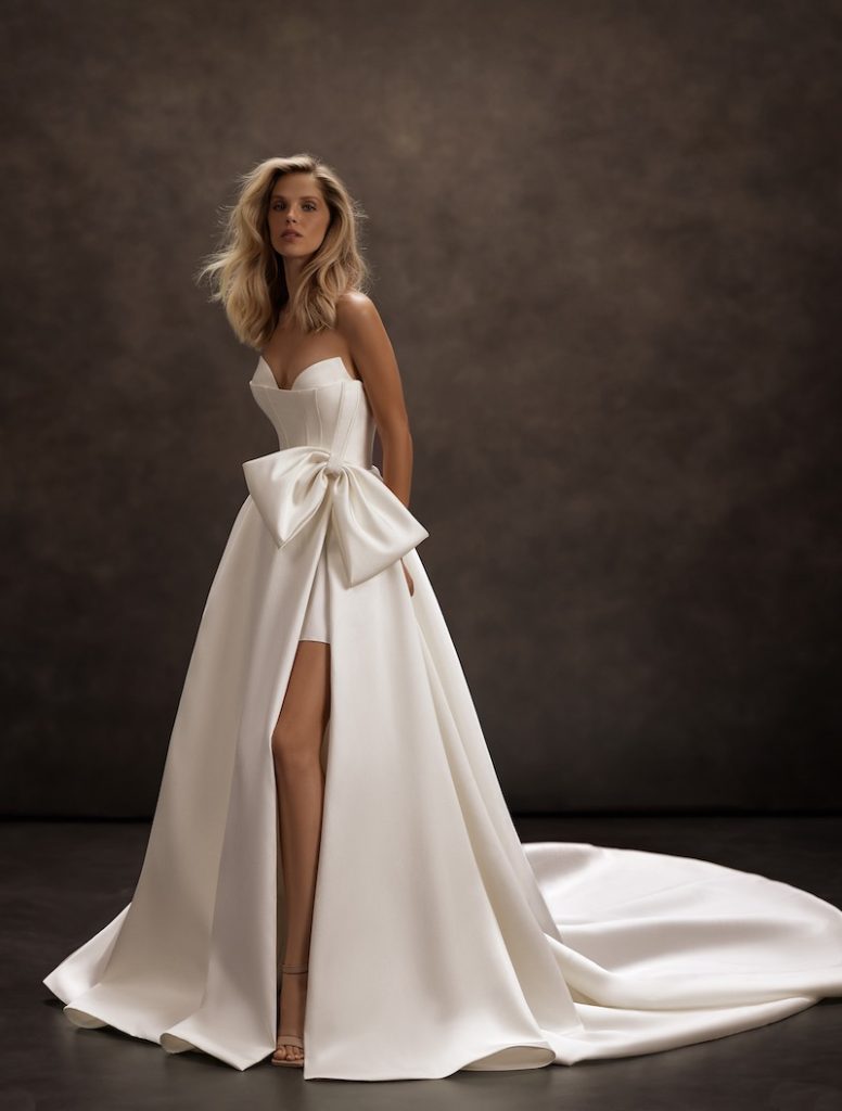 In questa foto, una modella posa  indossando un abito da sposa Jeorjett 2026 modello ad A in mikdao con spacco laterale, maxi fiocco, bustier 