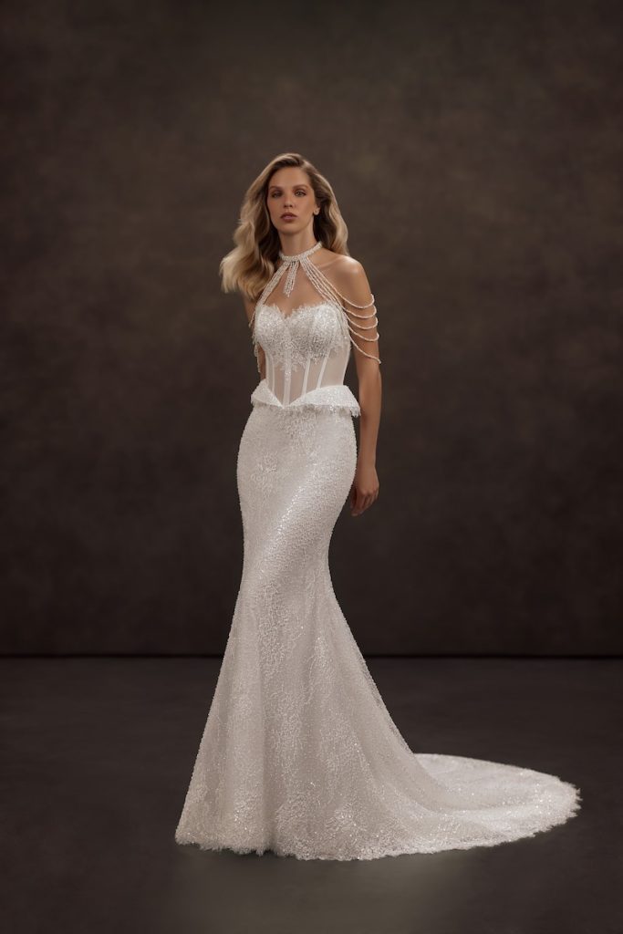 In questa foto, una modella posa  indossando un abito da sposa Jeorjett 2026 modello a sirena con ricami di microperline riprese anche sullo scollo che scende lungo le spalle 