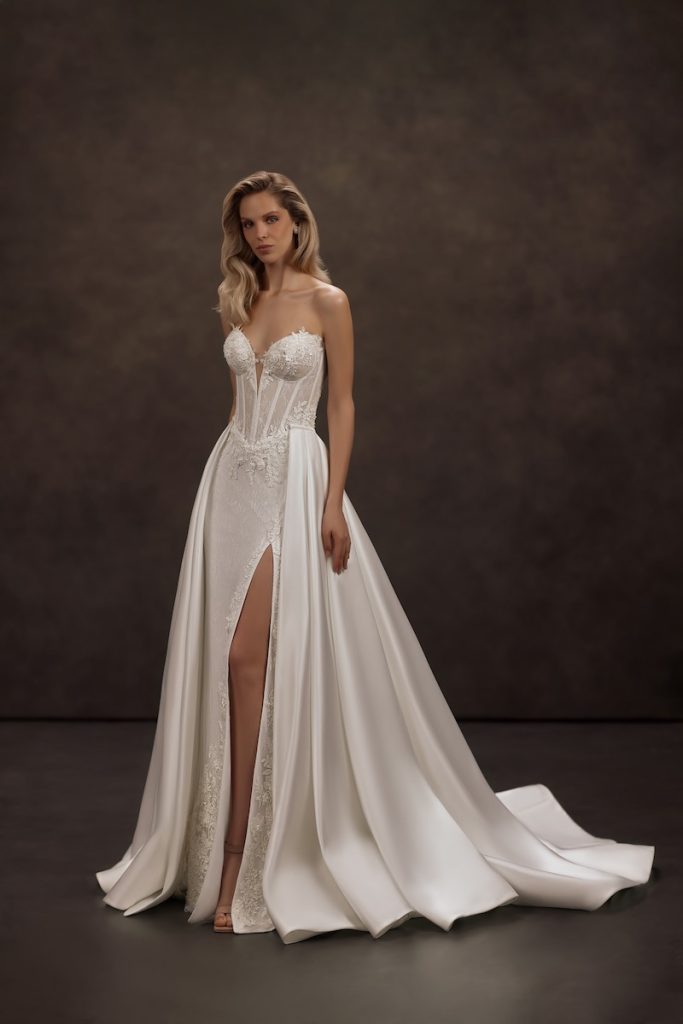 In questa foto, una modella posa  indossando un abito da sposa Jeorjett 2026 a sirena con riflessi preziosi, con spacco laterale, ricami floreali, scollo a cuore e sovragonna in mikado di seta