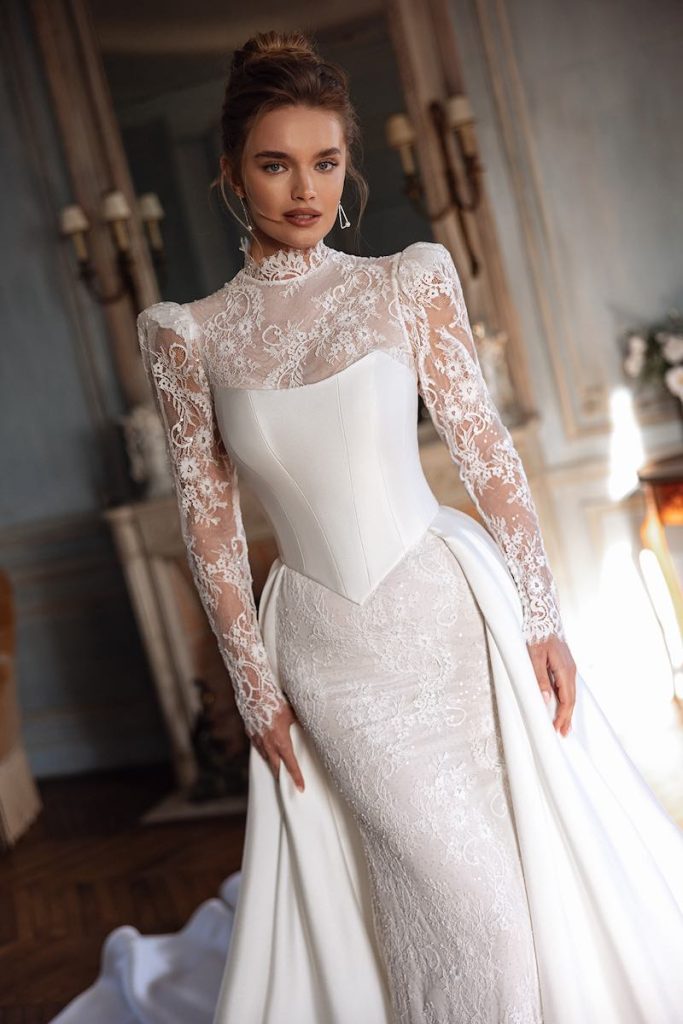In questa foto, una modella inquadrata a mezzo busto posa  indossando un abito da sposa Oliver Martino 2026 modello a sirena in pizzo con maniche lunghe, spalline alte, collo alto, con sovrapposti gonna e bustier steccato in mikado 