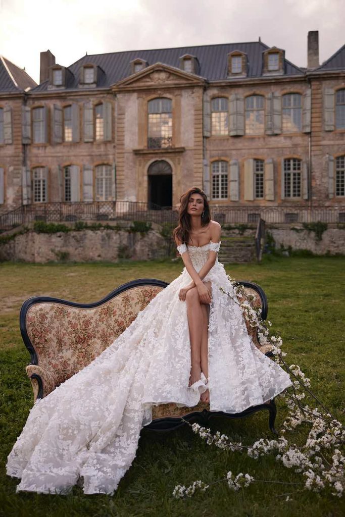 In questa foto, una modella posa seduta su un divano in un giardino di una dimora storica indossando un abito da sposa Oliver Martino 2026 in tulle ricamato con microfiori e spacco frontale