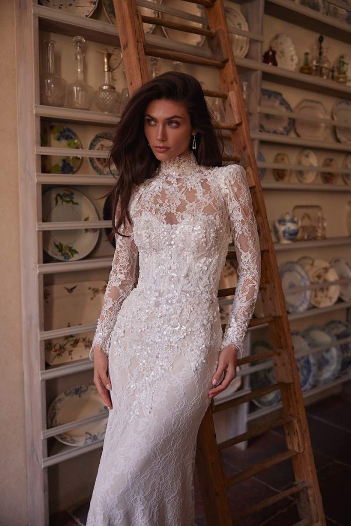In questa foto, una modella inquadrata a mezzo busto posa  indossando un abito da sposa Oliver Martino 2026 modello a sirena con maniche lunghe e collo alto, in pizzo decorato con perline e paillettes