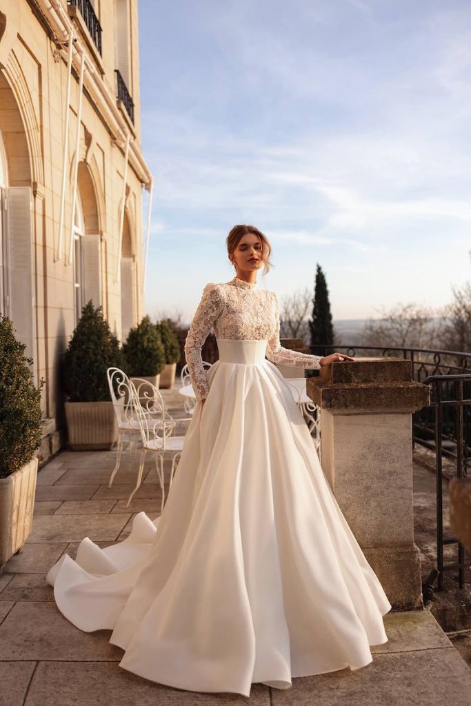In questa foto, una modella posa sul balcone di una dimora storica indossando un abito da sposa Oliver Martino 2026 con gonna ampia in mikado e corpetto in pizzo a collo alto e maniche lunghe