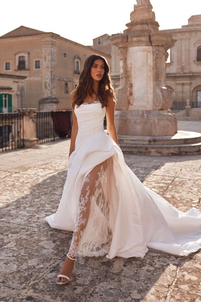 In questa foto, una modella posa  indossando un abito da sposa Pollardi 2026 in mikado con profondo spacco laterale decorato con tulle ricamato in pizzo 