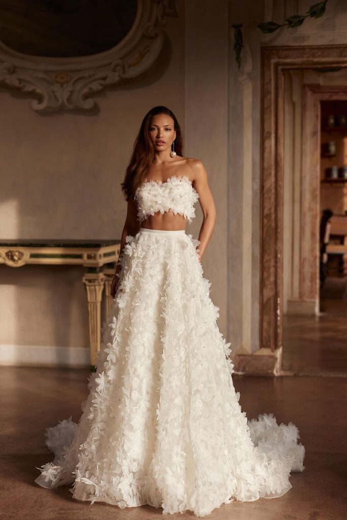In questa foto, una modella posa  indossando un abito da sposa Pollardi 2026 modello due pezzi con crop top e maxi gonna con fiori 3D all over