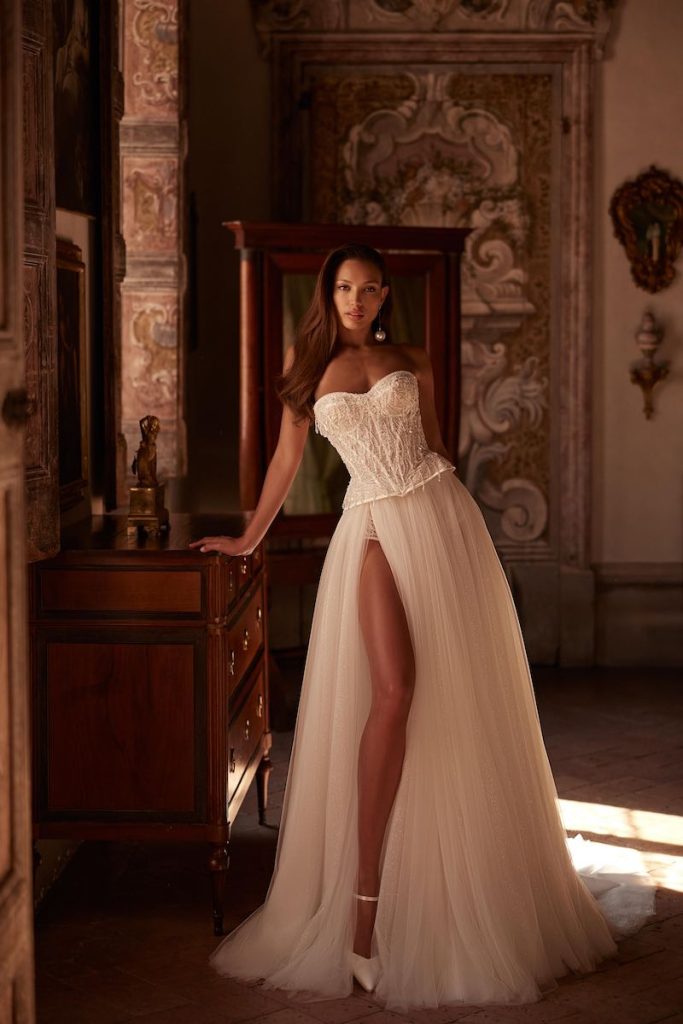 In questa foto, una modella posa  indossando un abito da sposa Pollardi 2026 modello scivolato con gonna in organza e bustier a vita basca con ricami di perline