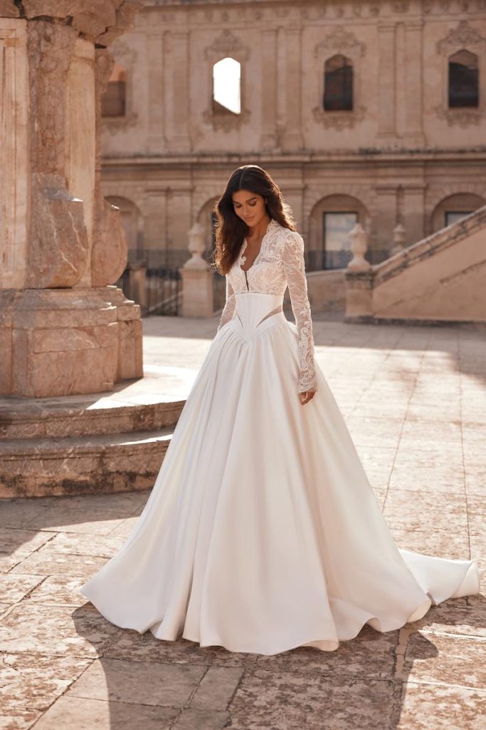 In questa foto, una modella posa  indossando un abito da sposa Pollardi 2026 modello ad A con gonna in mikado, maniche lunghe e scollo Queen Anne