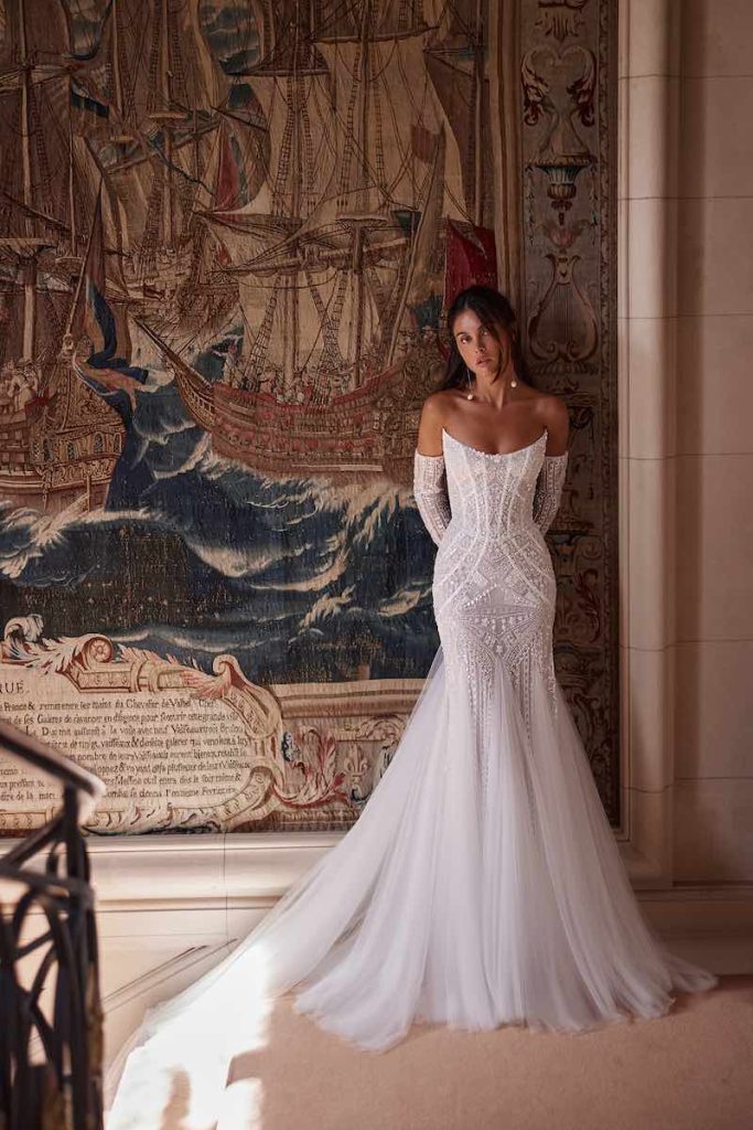 In questa foto, una modella posa  indossando un abito da sposa Yedyna 2026 modello a semi sirena con ricami, strascico in tulle e manicotti abbinati