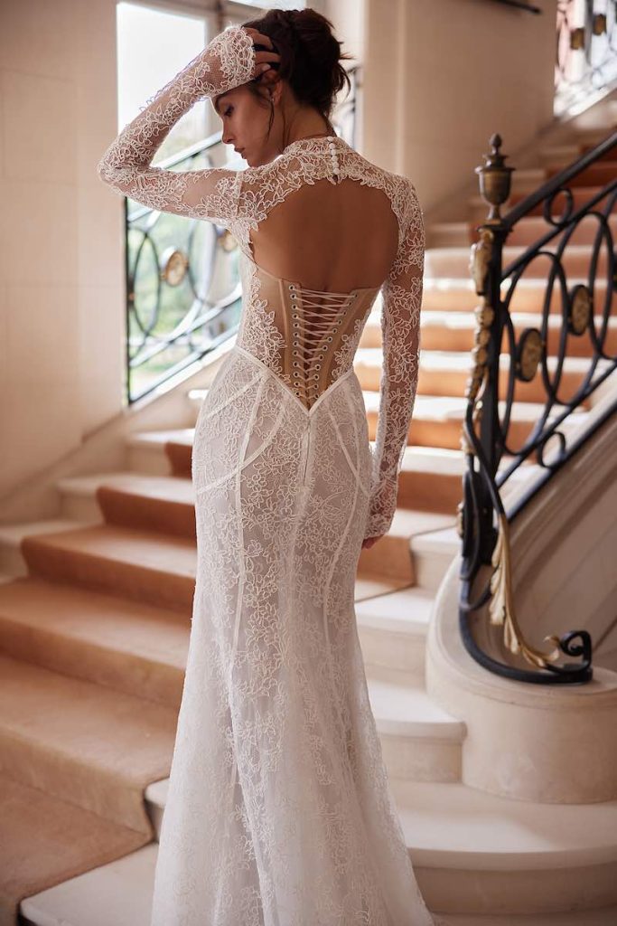 In questa foto, una modella posa di spalle indossando un abito da sposa Yedyna 2026 modello a sirena con bustier stringato a vista e coprispalle in pizzo che lascia la scollatura ad oblò sulla schiena