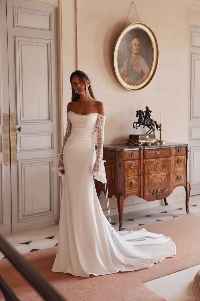 In questa foto, una modella posa di spalle indossando un abito da sposa Yedyna 2026 modello a semisirena in tessuto stretch con corpetto drappeggiato e maniche a campana in pizzo strette al polso con un microfiocchi