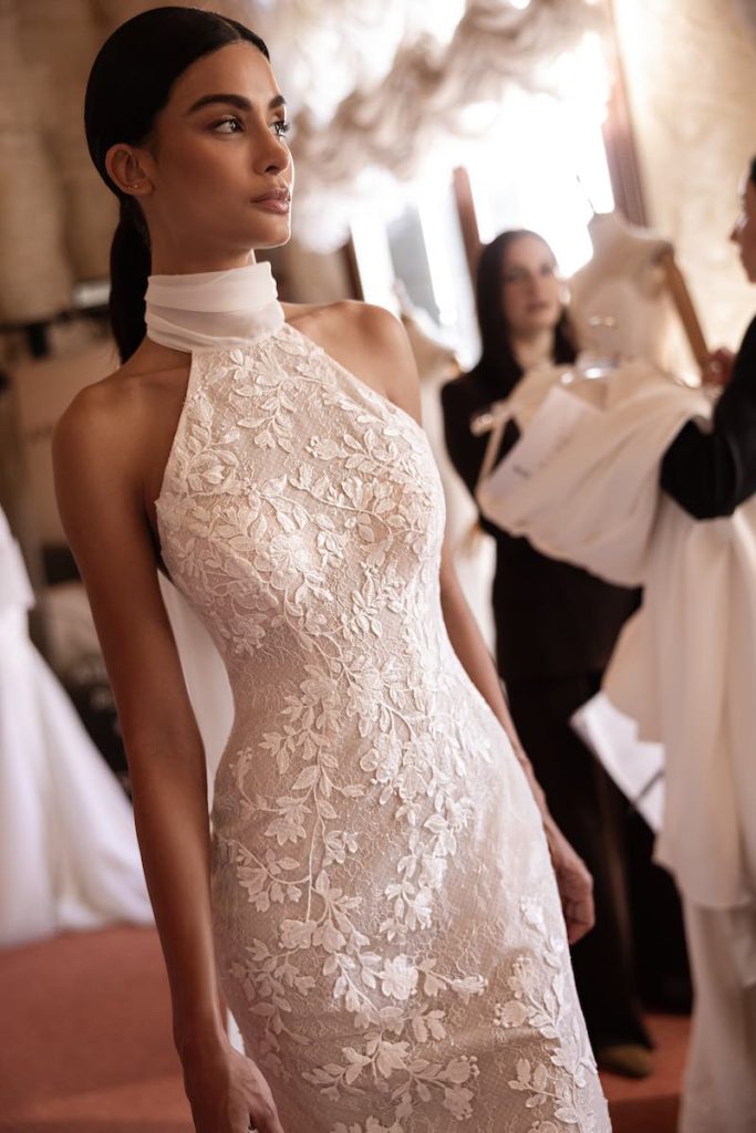 In questa foto, una modella posa indossando un abito da sposa Anna Mariee 2026 a sirena in pizzo floreale con scollo halter 