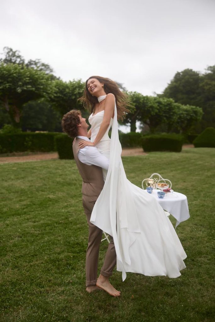 In questa foto, una modella posa con un modello che la solleva in alto roteando sul prato, indossando un abito da sposa Anna Mariee 2026 a sirena con lungo strascico e un lungo foulard in chiffon al collo