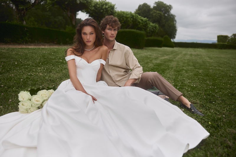 In questa foto, una modella posa con un modello, entrambi seduti su un prato, indossando un abito da sposa Anna Mariee 2026 modello ampio con scollo off shoulder