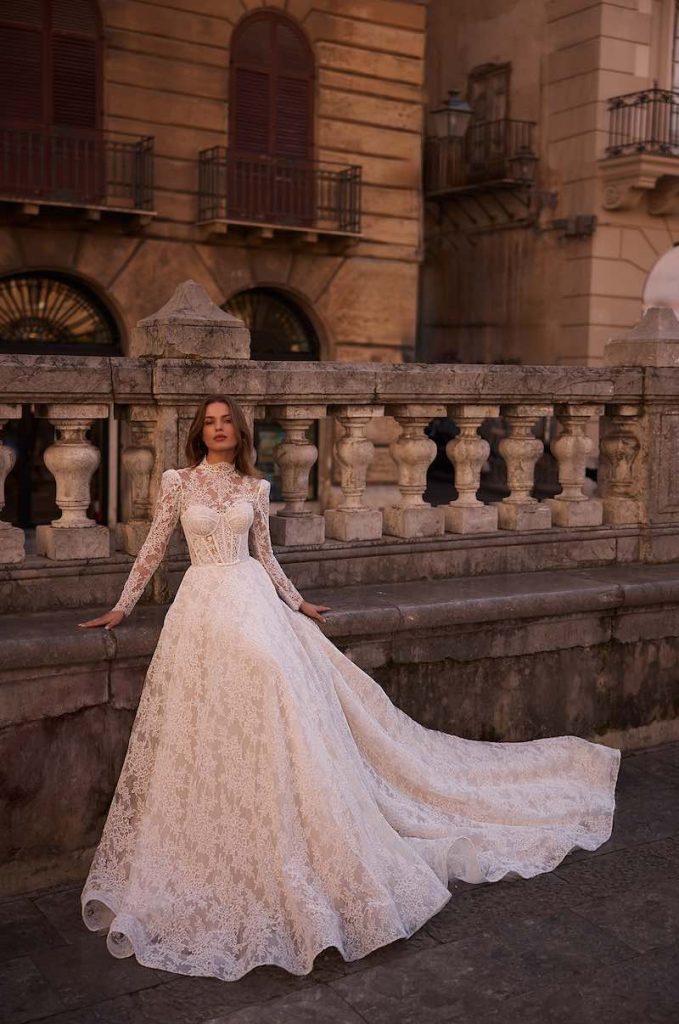 In questa foto, una modella posa indossando un abito da sposa Aria 2026 dal modello ad A interamente in pizzo con maniche lunghe e a collo alto con bustier steccato a vista 