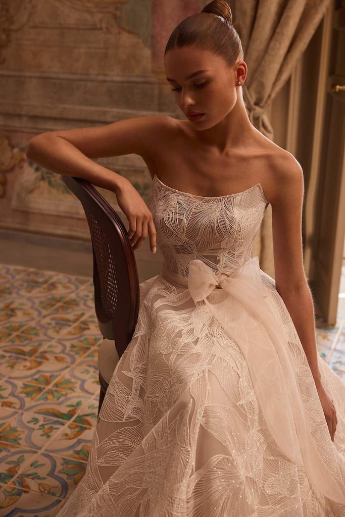 In questa foto, una modella posa indossando un abito da sposa Aria 2026 con scollatura dritta, fiocco in vita e organza ricamata con motivi floreali 