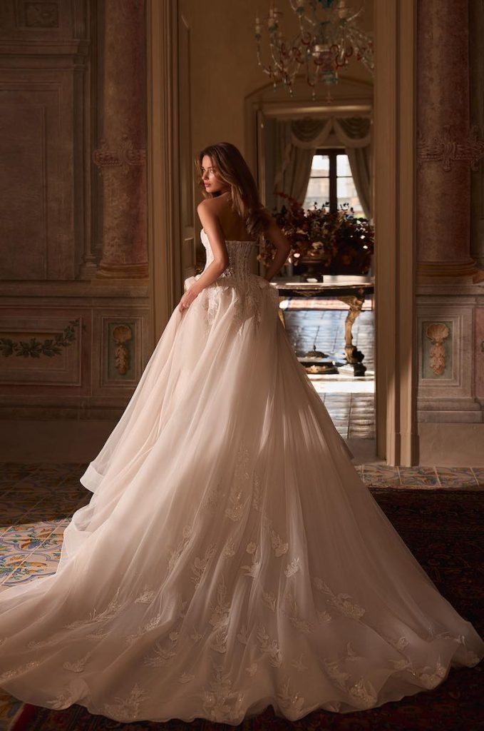 In questa foto, una modella posa di spalle indossando un abito da sposa Aria 2026 con gonna ad A e lungo strascico decorato con fiori 3D
