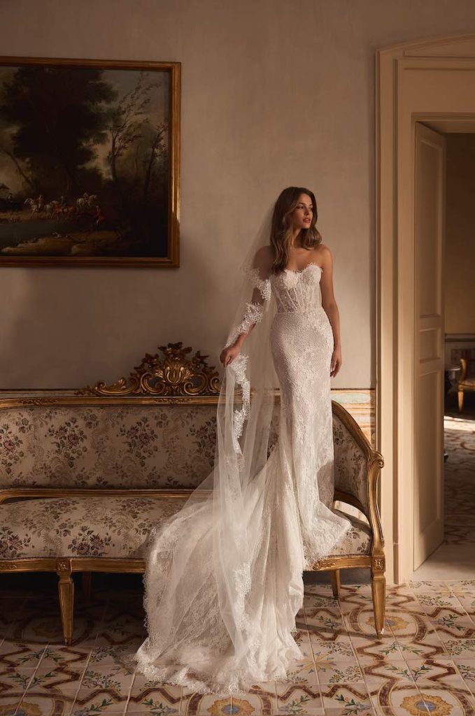 In questa foto, una modella posa su un divanetto stile ottocentesco con tappezzeria floreale indossando un abito da sposa Aria 2026 modello a sirena in oizzo con corpetto steccato, scollo a cuore, lungo strascico e velo abbinato con ricami sul bordo 