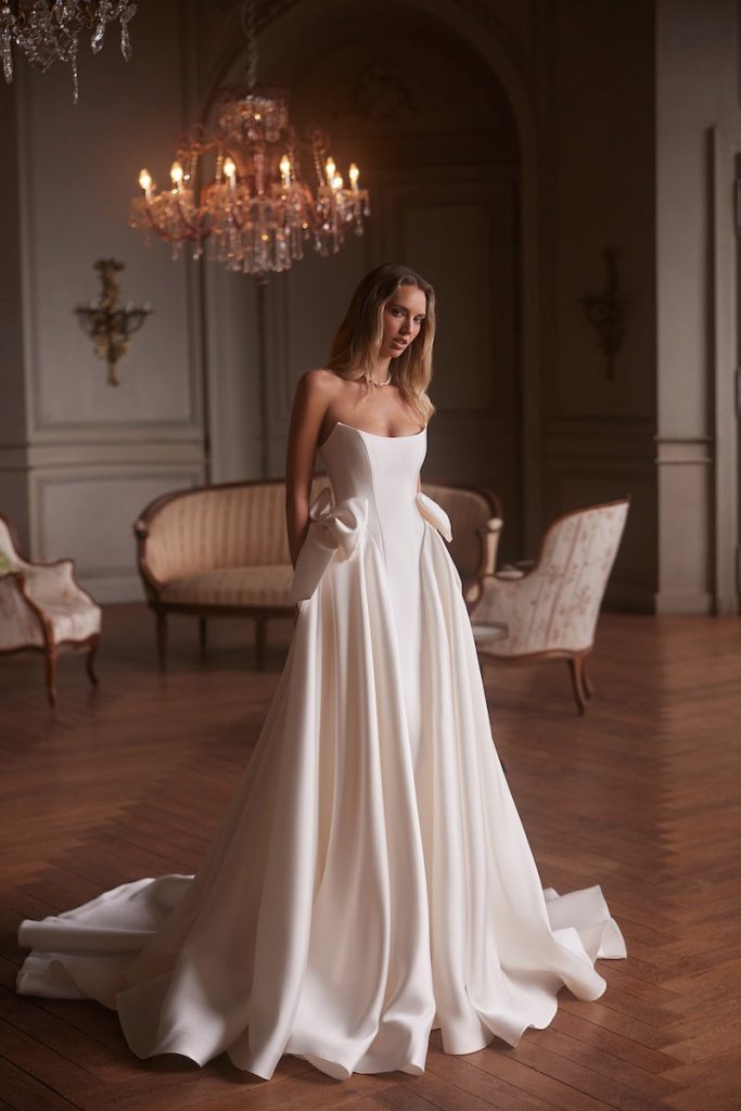 In questa foto, una modella posa indossando un abito da sposa Armonia 2026 in mikado di seta con drappeggio laterale e scollatura dritta