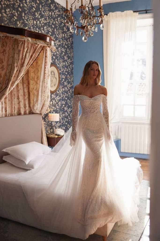 In questa foto, una modella posa indossando un abito da sposa Armonia 2026 modello a sirena con maniche lunghe, scollo off shoulder e sovragonna in tulle 