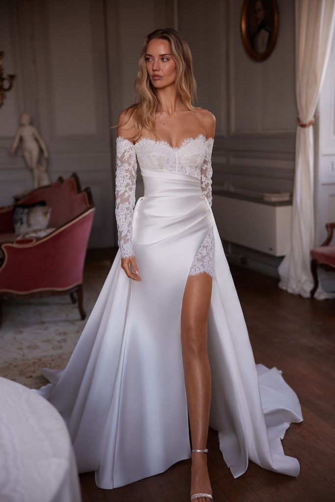 In questa foto, una modella posa indossando un abito da sposa Armonia 2026 in pizzo, con manicotti abbinati , scollo a cuore e sovragonna in mikado di seta 