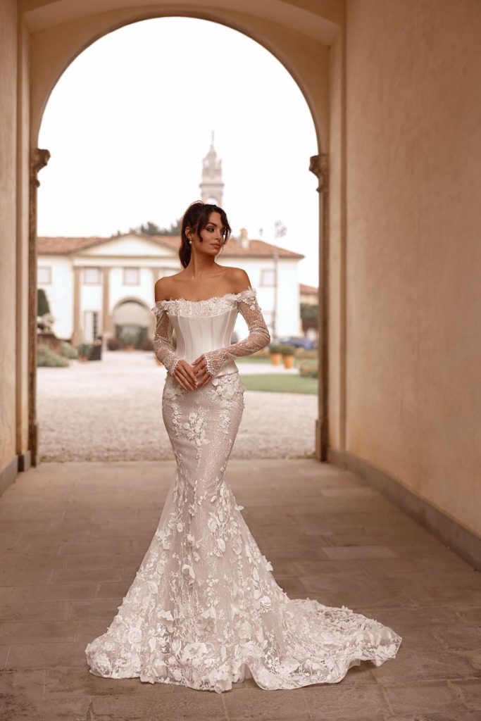 In questa foto, una modella posa indossando un abito da sposa Dominiss 2026 modello semi sirena in pizzo con ricami 3D e bustier staccato e steccato a vita basca con scollo off shoulder e maniche lunghe 