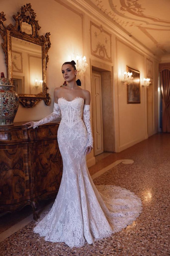 In questa foto, una modella posa indossando un abito da sposa Dominiss 2026 modello a semi sirena, interamente in pizzo, con bustier steccato, scollo a cuore e manicotti abbinato