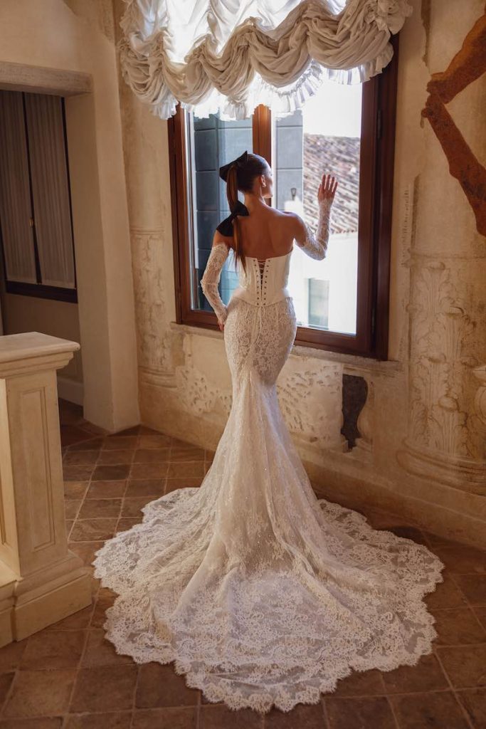In questa foto, una modella posa di spalle indossando un abito da sposa Dominiss 2026 modello a sirena, in pizzo, con manicotti abbinati e bustier con chiusura a vista 