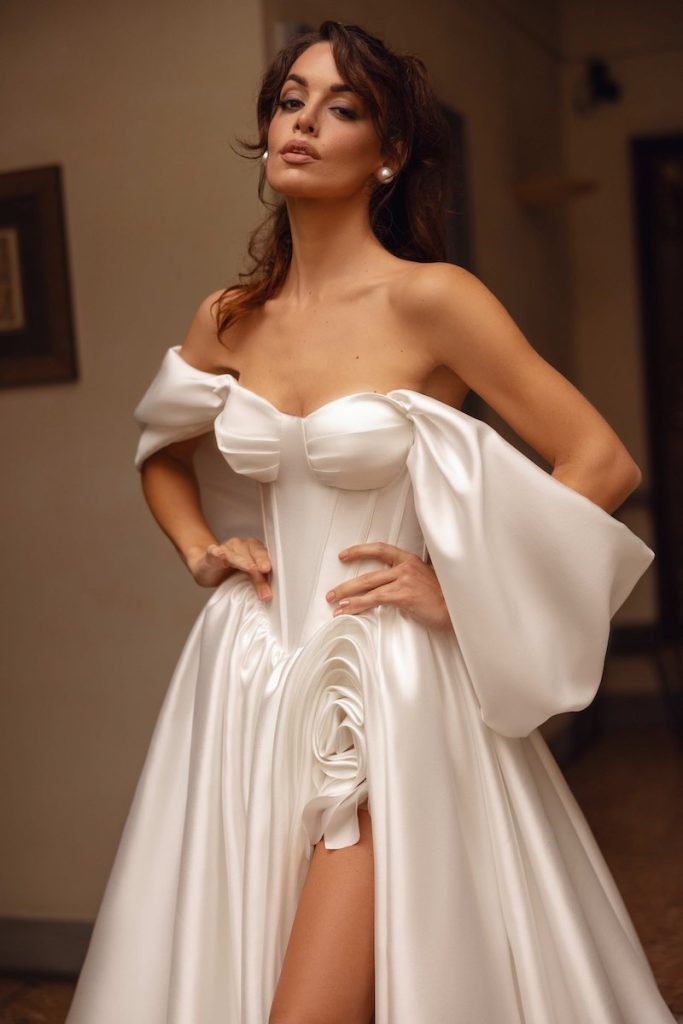 In questa foto, una modella inquadrata a mezzo busto posa indossando un abito da sposa Dominiss 2026 con bustier steccato in stile elisabettiano, scollo a cuore, maxi maniche e decoro di una maxi rosa sullo spacco