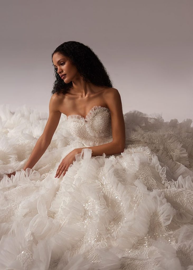 In questa foto, una modella posa seduta indossando un abito da sposa Elena Morar 2026 con ruches all over 
