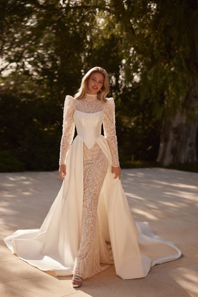 In questa foto, una modella posa  indossando un abito da sposa Elena Morar 2026 modello a sirena in pizzo con maniche lunghe, spalline alte, collo alto, con sovrapposti gonna e bustier steccato in mikado 