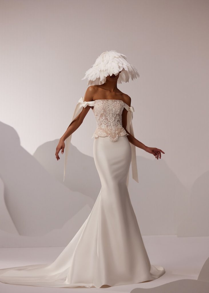 In questa foto, una modella posa indossando un abito da sposa Elena Morar 2026 modello a semi sirena con gonna in mikado e corpetto sovrapposto in pizzo con fiocchi sulle braccia. Il look è completato da un cappello di pium